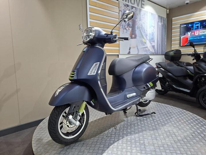 A sleek blue vintage scooter parked on a metallic display stand, with modern design elements and a stylish profile, set against a backdrop featuring a cityscape and a television screen.