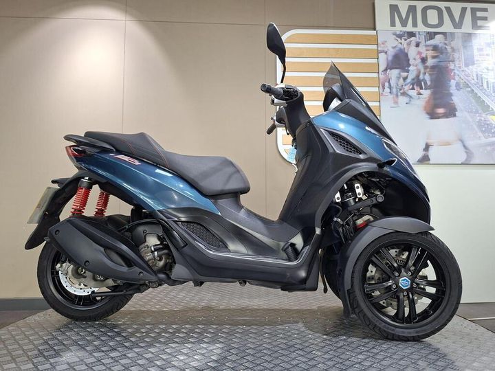 Piaggio MP3 300 Sport 2024 A blue three-wheeled scooter parked on a textured floor, showcasing a sleek design with red shock absorbers and a black seat, against a light beige wall with a blurred background image of people walking.