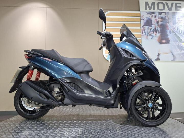 Piaggio MP3 300 Sport 2024 A blue three-wheeled scooter parked on a textured floor, showcasing a sleek design with red shock absorbers and a black seat, against a light beige wall with a blurred background image of people walking.