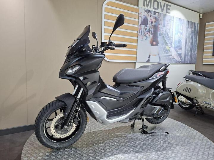 A black and silver scooter displayed in a showroom with a minimalist design, featuring a prominent front fairing, large wheels, and a digital display, against a beige background and a wall graphic depicting an urban scene.
