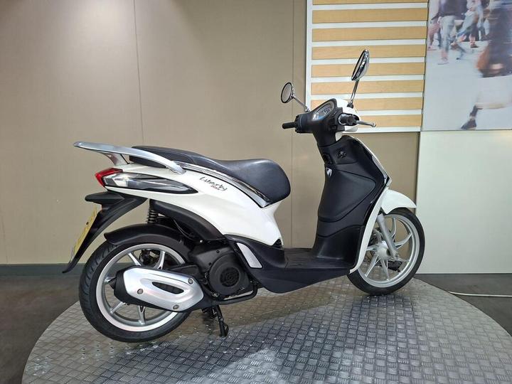 A white scooter displayed on a circular platform, showcasing its side profile. The design features a sleek body with a black seat, silver wheels, and rearview mirrors. The background has a neutral tone, highlighting the scooter's details.