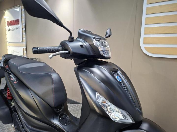 A close-up view of a sleek black scooter, highlighting the handlebars, mirror, and part of the seat. The scooter features modern design elements and is displayed against a neutral background with branding visible.
