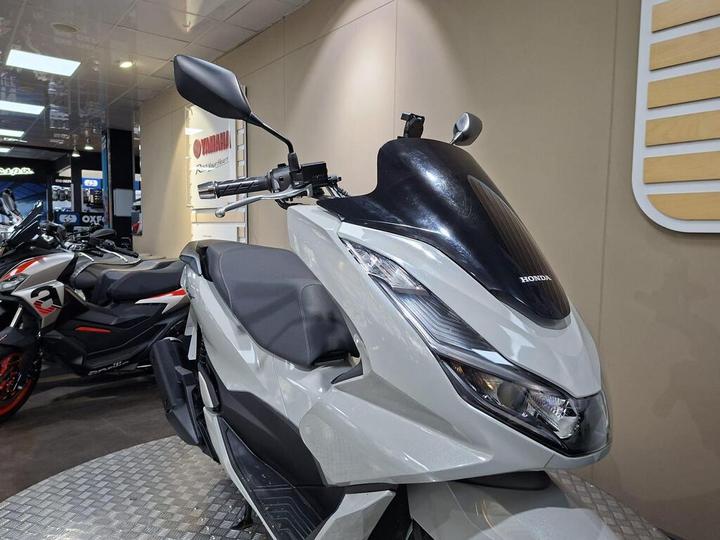 A close-up view of a white Honda scooter with a sleek design, including an aerodynamic windshield and handlebars, displayed in a modern showroom with other motorcycles in the background.