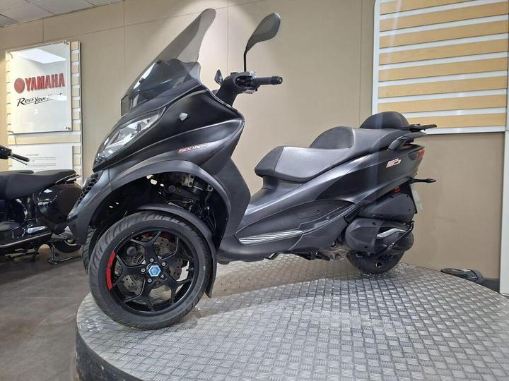 A sleek black three-wheeled scooter displayed on a round, textured platform with a Yamaha sign in the background.