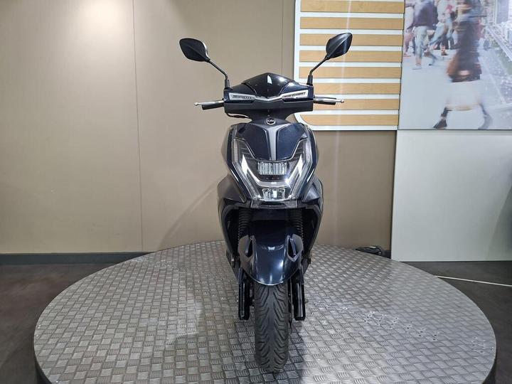 A sleek, modern scooter displayed front-facing on a circular platform, with a shiny black body, distinctive headlights, and mirrors, set against a neutral background.