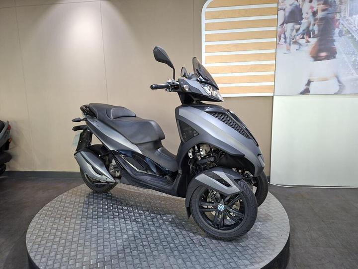 A sleek, gray three-wheeled scooter displayed on a circular platform with a textured surface, set against a neutral wall.