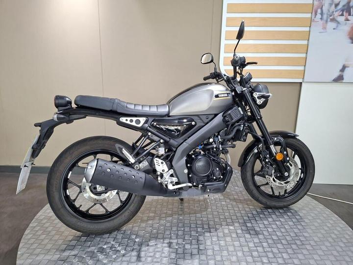 A side view of a Yamaha motorcycle in a showroom, featuring a sleek gray frame, black accents, and a sporty design, displayed on a textured platform against a minimalistic backdrop.