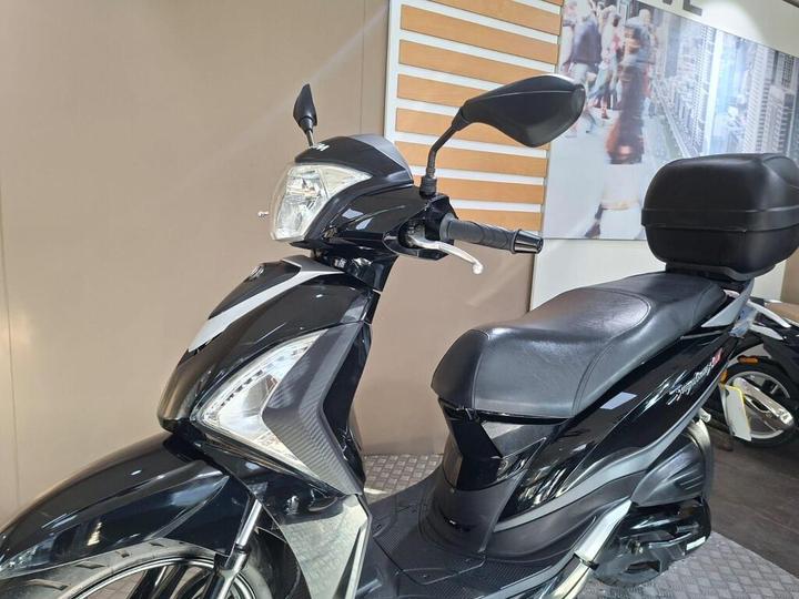 A sleek black scooter with a streamlined design, showcasing its front headlight, side mirror, and comfortable seat, set against a light-colored wall with a blurred background of a city scene.