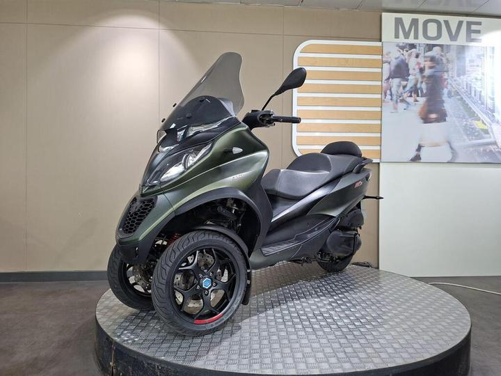 A green three-wheeled scooter displayed on a circular platform with a textured surface, featuring a transparent windshield, sleek design, and sporty black wheels. The background includes promotional signage and a blurred image of people walking.