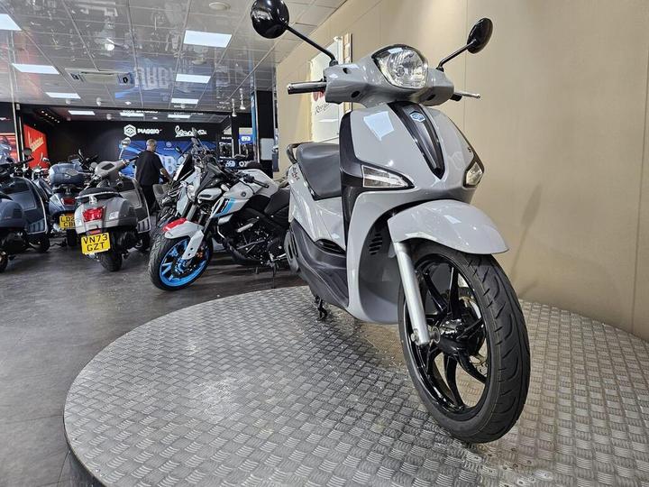 A gray scooter is prominently displayed on a circular platform in a motorcycle showroom, with various other motorcycles visible in the background. A person is examining one of the bikes.