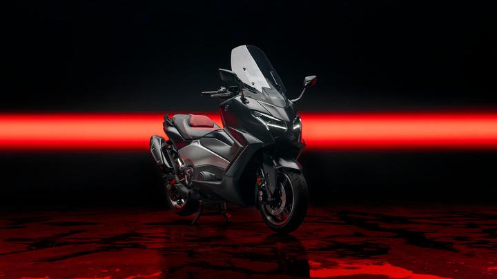 A sleek, modern motorcycle with a dark metallic finish is positioned against a black background, accented by a striking red light strip. The bike features a tall windshield, stylish body lines, and a comfortable seat with red details. The reflective surface below enhances the dramatic effect of the scene.