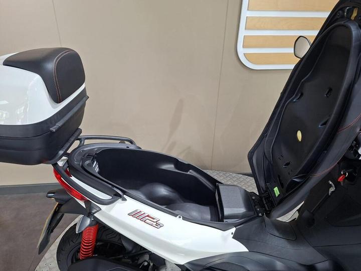 A close-up view of an open motorcycle storage compartment with a sleek, modern design, featuring a black and white exterior and red accents. The compartment is spacious, revealing a smooth interior, and is positioned next to a tan wall.