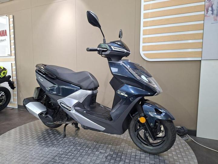 A sleek blue and silver scooter displayed on a textured floor, featuring a low-profile design and modern handlebar controls, with a minimalist background.