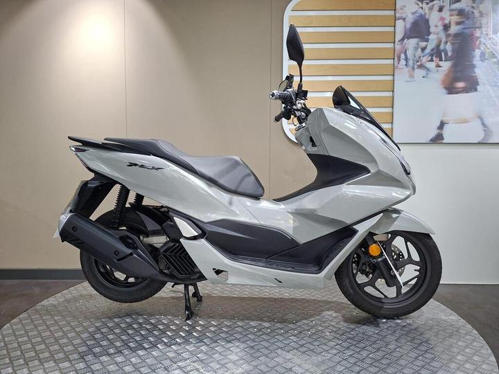 Honda PCX 125 (2024) A sleek gray scooter displayed on a circular platform with a textured surface, showcasing its modern design and prominent features, set against a neutral wall background.
