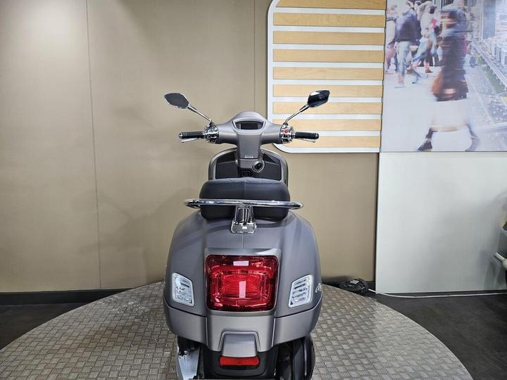 A modern gray scooter is displayed from the rear, showcasing its sleek design and chrome details, in a showroom setting with a blurred image of pedestrians in the background.