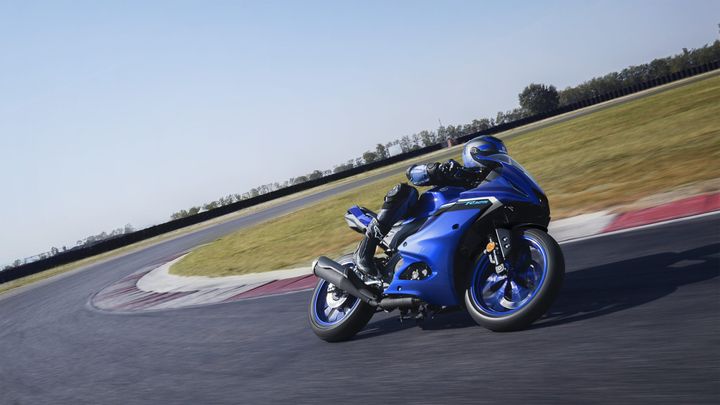 A blue motorcycle leans into a curve on a racetrack, showcasing dynamic motion against a clear sky and green grass.