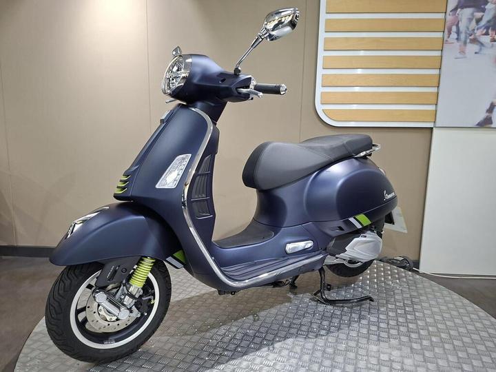A sleek blue scooter with green accents is displayed on a textured surface, showcasing its modern design and chrome details against a neutral background.