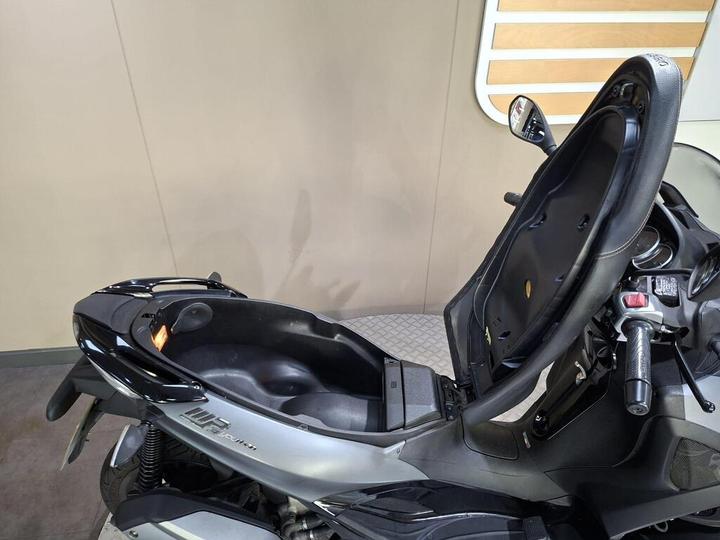 A close-up view of a black and silver scooter with the seat lifted, revealing storage space underneath, set against a neutral background.