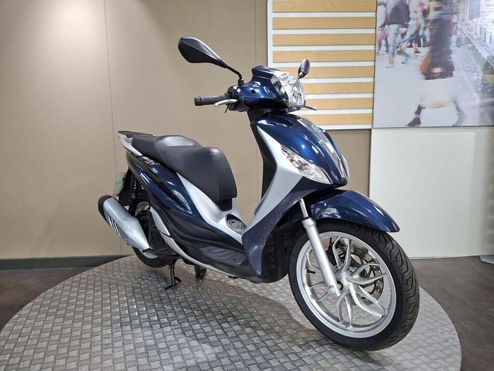 A blue and silver scooter is displayed on a textured circular platform against a plain background with a large image of people walking in the upper corner.
