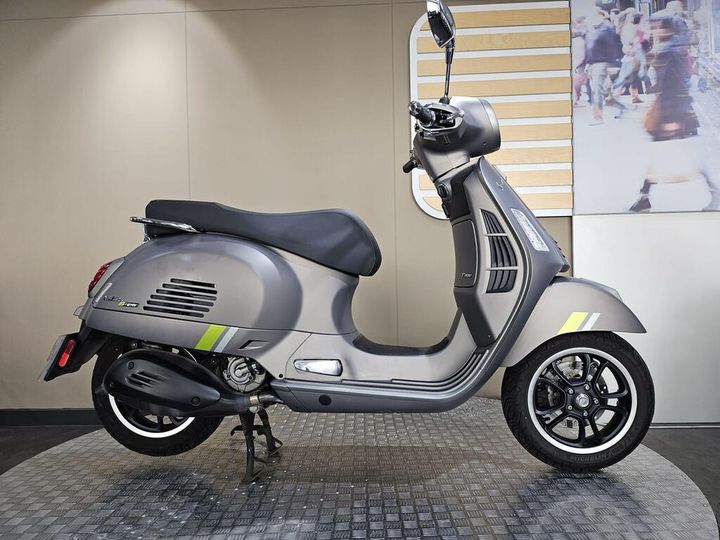 Vespa GTS 300 SuperTech (2023) A sleek gray scooter showcased against a neutral background, featuring a comfortable seat and modern design elements, with prominent yellow accents on the body and a stylish wheel.