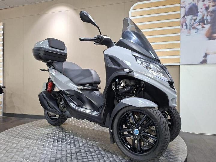 A sleek grey three-wheeled scooter displayed on a circular metal platform in a modern showroom, featuring a black seat, storage compartment on the back, and red suspension accents.
