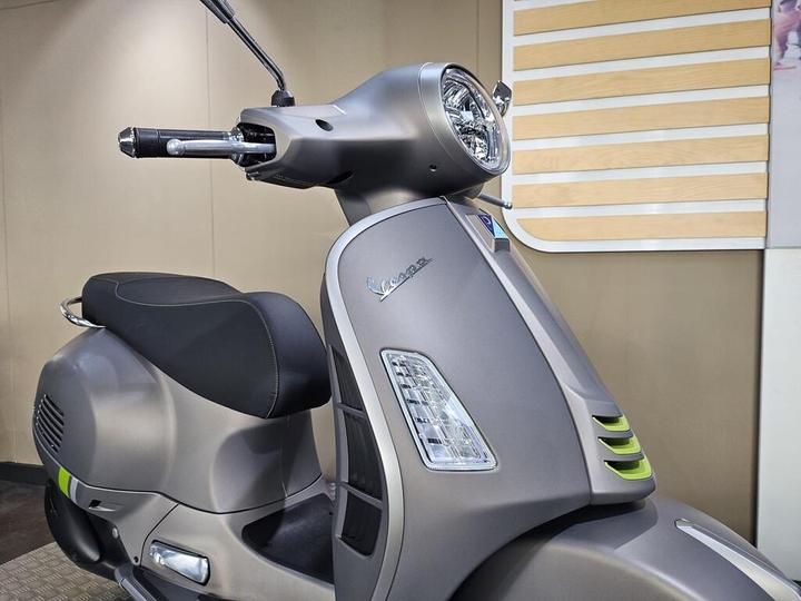 A close-up view of a modern gray scooter parked indoors, showcasing its front design, headlight, and handlebar with grips, against a neutral background.