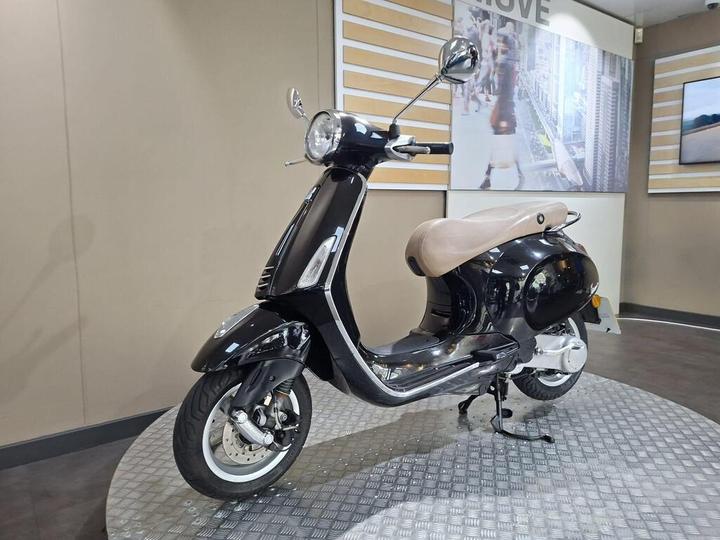 A sleek black scooter with a beige seat displayed on a textured platform in a showroom, with a modern interior in the background featuring wall graphics.