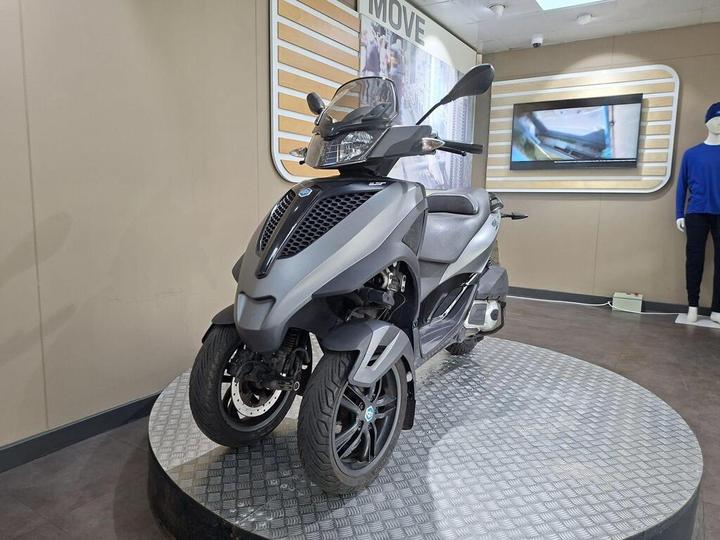 A gray three-wheeled scooter displayed on a circular platform in an indoor showroom, with a modern interior design featuring wall panels and a large screen in the background.