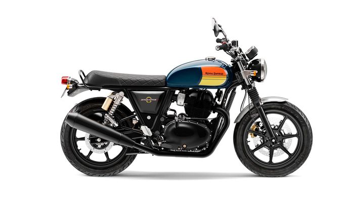 Alt text: A Royal Enfield Interceptor motorcycle in a studio setting, featuring a black frame, blue and orange fuel tank, and a diamond-stitched black seat.