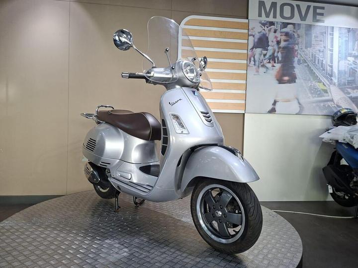 A silver Vespa scooter displayed on a circular platform, with a brown seat and a transparent windshield, set against a minimalistic background featuring a "MOVE" sign and an image of a bustling street.