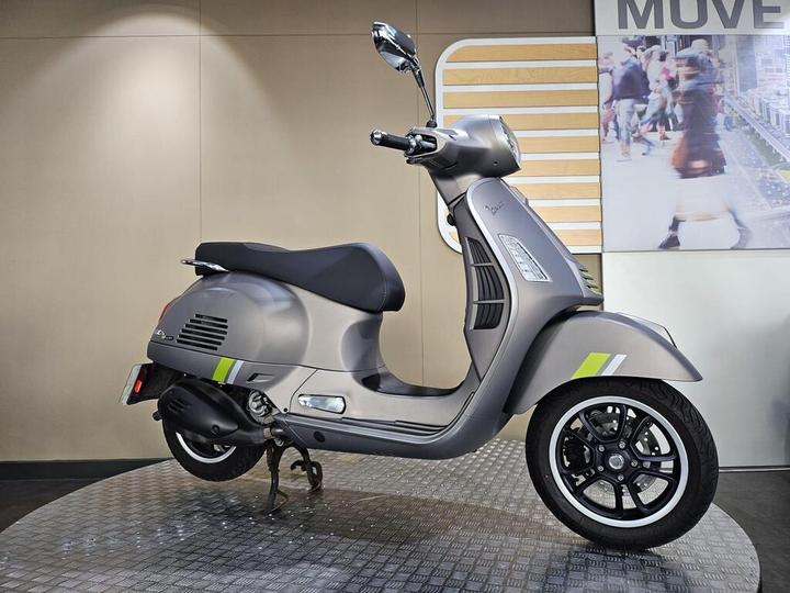 A sleek, gray scooter stands on a textured platform, showcasing its modern design with white and green accents. The scooter features a rounded headlight, a comfortable seat, and stylish black wheels, set against a minimalist background with a blurred urban scene.