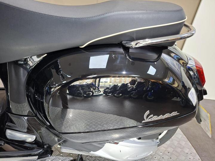 Close-up of a black Vespa scooter's side panel, featuring a shiny reflective surface and a visible logo, with a blurred background of more scooters.