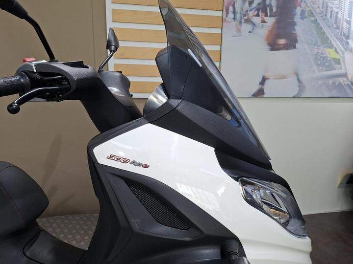 Close-up of a white scooter with a prominent "300" badge, featuring a sleek design, an adjustable windshield, and handlebars in a showroom setting.