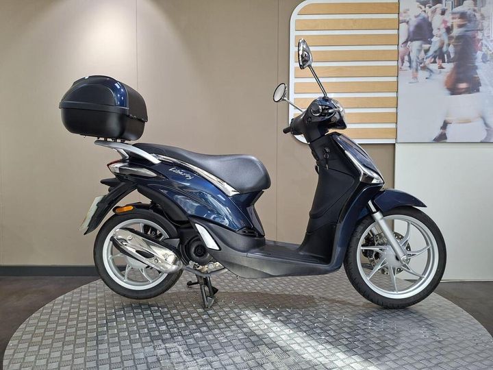 Piaggio Liberty 50 (2019) A blue scooter with a black top case is displayed on a circular textured base, set against a neutral background.