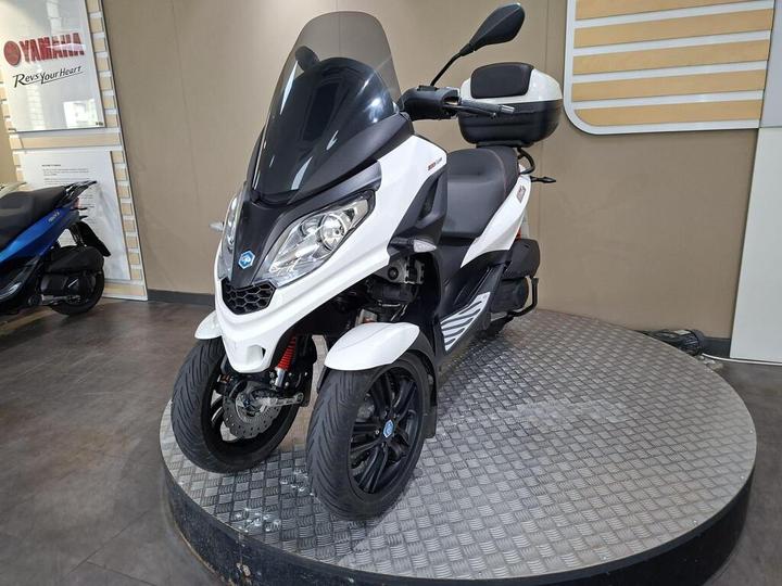 A white and black three-wheeled scooter is displayed on a circular platform inside a showroom, with a Yamaha logo visible in the background. The scooter features a large windshield, a comfortable seat, and a storage compartment at the back.
