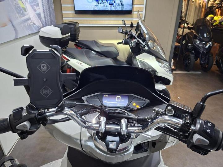 A close-up view of a scooter's handlebars and dashboard, showcasing a digital speedometer displaying 0 mph, multifunction controls, and a phone holder. In the background, several scooters and motorcycles are visible.