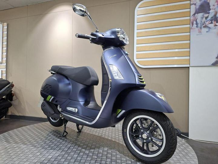 A blue Vespa scooter displayed in a showroom, featuring a sleek design with black seating and stylish alloy wheels, set against a modern, neutral-colored wall.