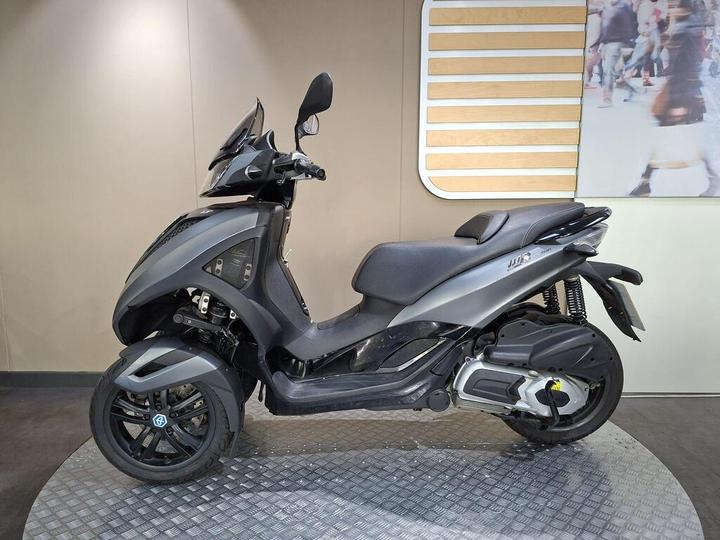 A sleek black three-wheeled scooter displayed on a circular platform, set against a light-colored wall with a modern background featuring people in motion.