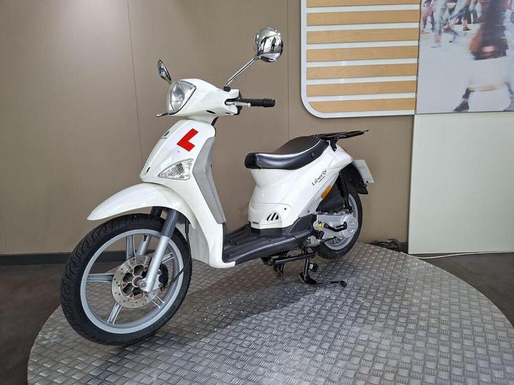 A white scooter with a red 'L' plate displayed on a circular metal platform in a modern indoor setting.