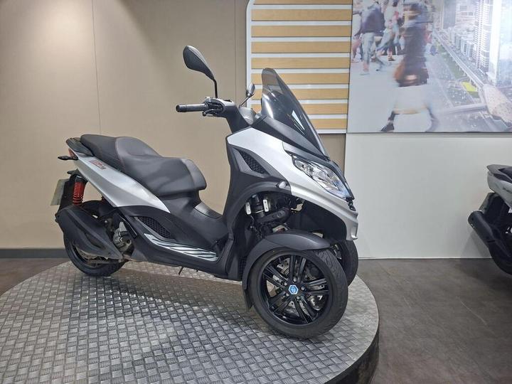 A three-wheeled scooter in silver and black, displayed on a circular platform against a light-colored wall, with a blurred background showing urban scenery.