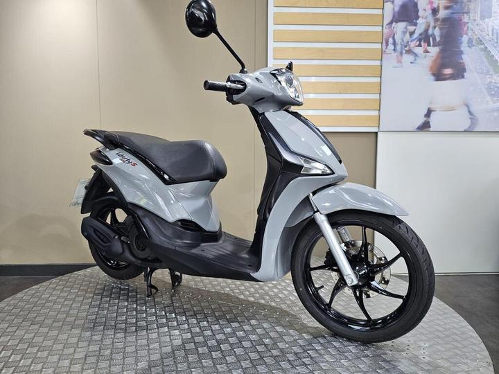 A gray scooter is displayed on a circular metallic platform in a showroom, with a minimalist beige wall and a blurred image of a city in the background.