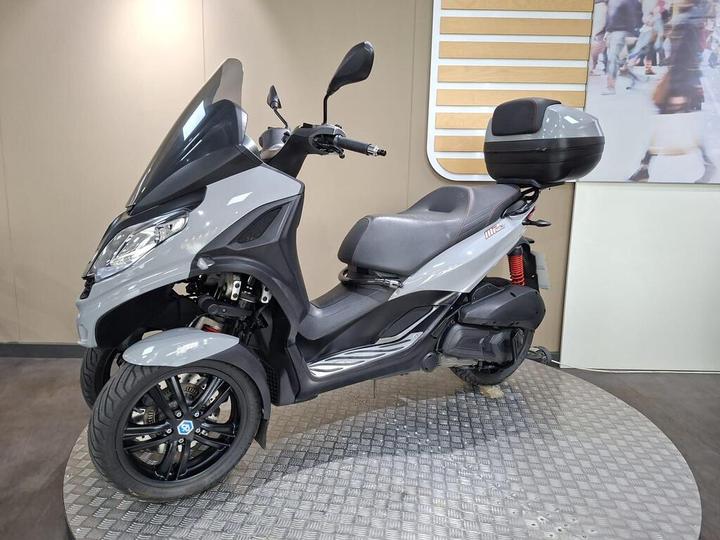 A gray three-wheeled scooter displayed on a circular platform, featuring a large front windscreen, comfortable seating with a backrest, and a rear storage compartment. The background is a neutral-colored wall with a blurred image of pedestrians.