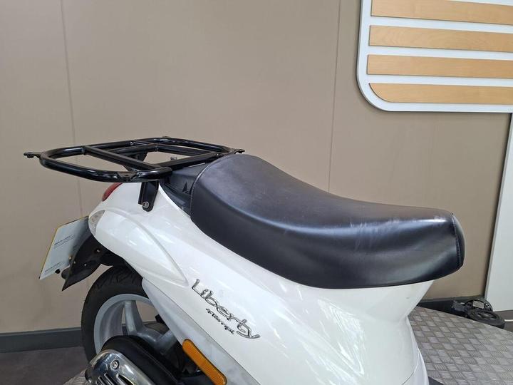 A close-up view of the rear section of a white scooter featuring a black seat and a metal luggage rack on top, set against a neutral-colored background.