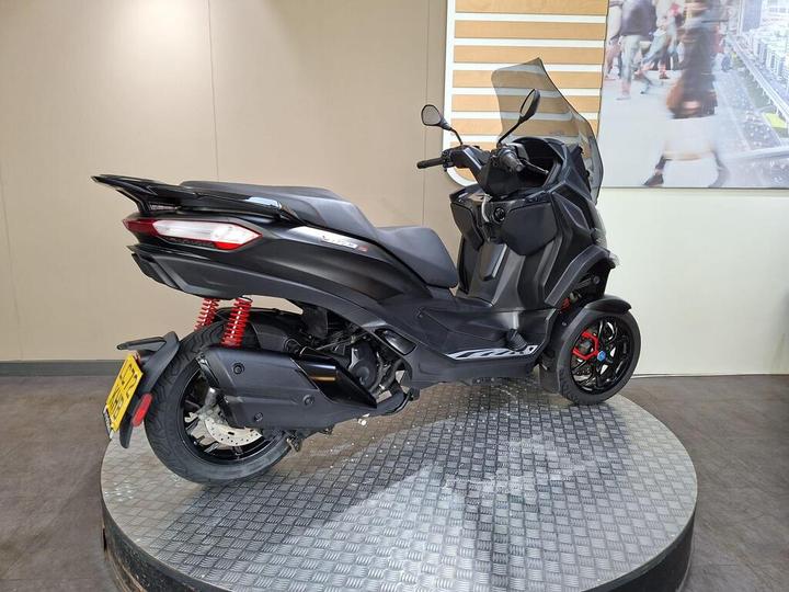 A black three-wheeled scooter with red suspension springs, displayed on a circular platform with a textured surface, against a neutral background.