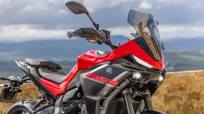 Close-up of a red and black Yamaha motorcycle, featuring a sleek design, prominent headlights, and a tall windscreen, set against a mountainous landscape under a cloudy sky.