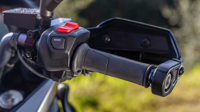 Close-up of a motorcycle handlebar grip featuring a MODE button and a textured throttle grip against a blurred natural background.