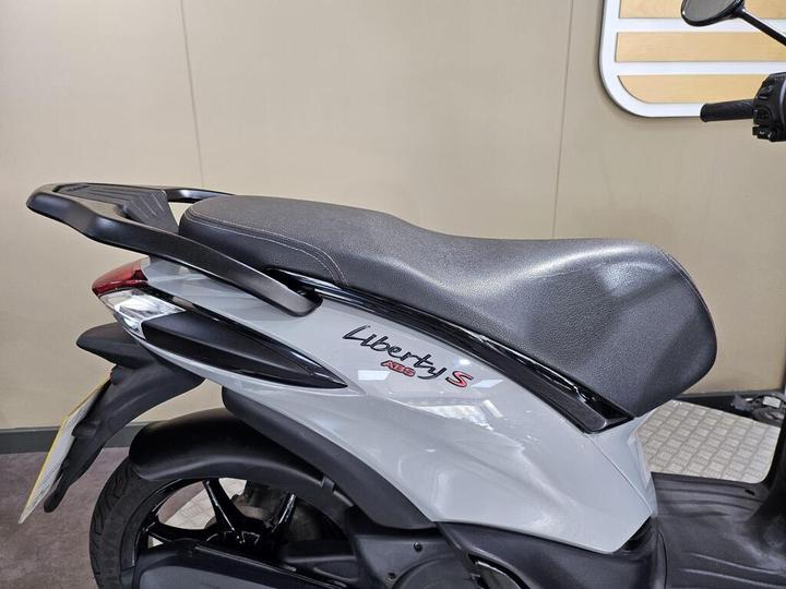 A close-up view of the rear section of a gray scooter, featuring the "Liberty S" logo on the side, with a textured black seat and sleek design elements.