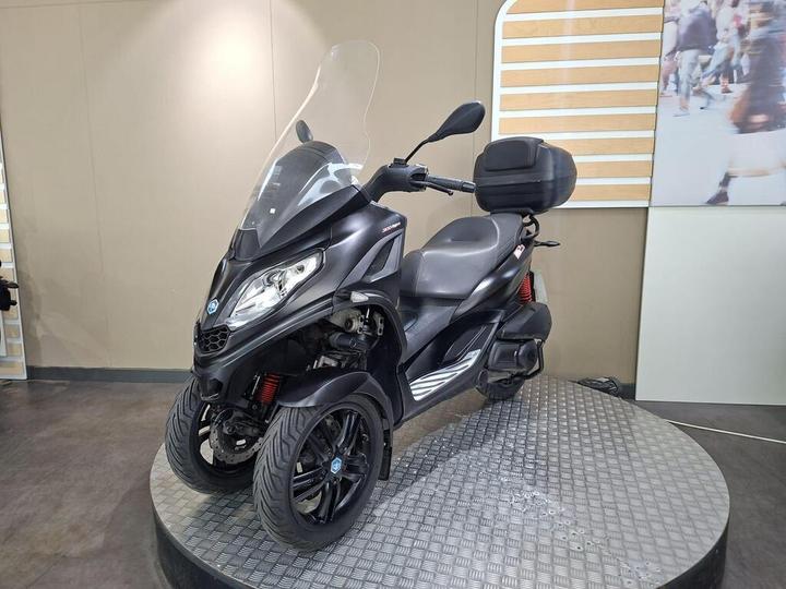 A black three-wheeled scooter with a windscreen and a small rear storage compartment, displayed on a circular platform in a contemporary setting.
