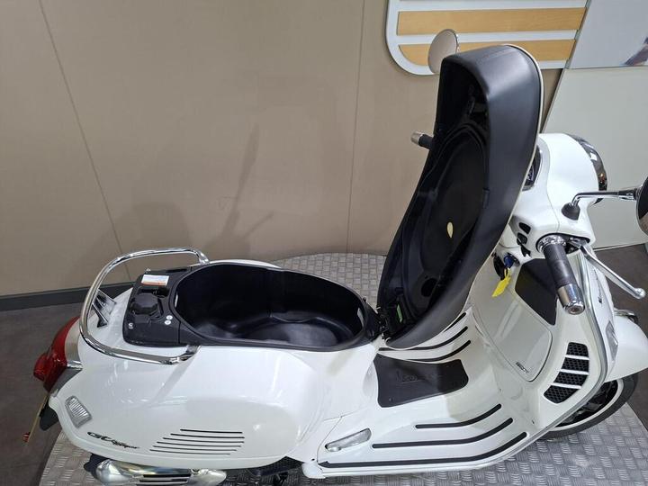 A white scooter with a raised seat, revealing a spacious storage compartment beneath. The scooter has a streamlined design, chrome accents, and is displayed on a textured surface against a neutral background.