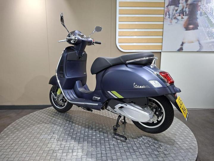 A stylish blue Vespa scooter is displayed on a circular stand, showcasing its distinctive design, including a sleek body, comfortable seat, and chrome accents, set against a neutral background.